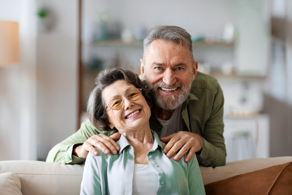 A smiling older couple at home, with the man resting his hands on the woman's shoulders