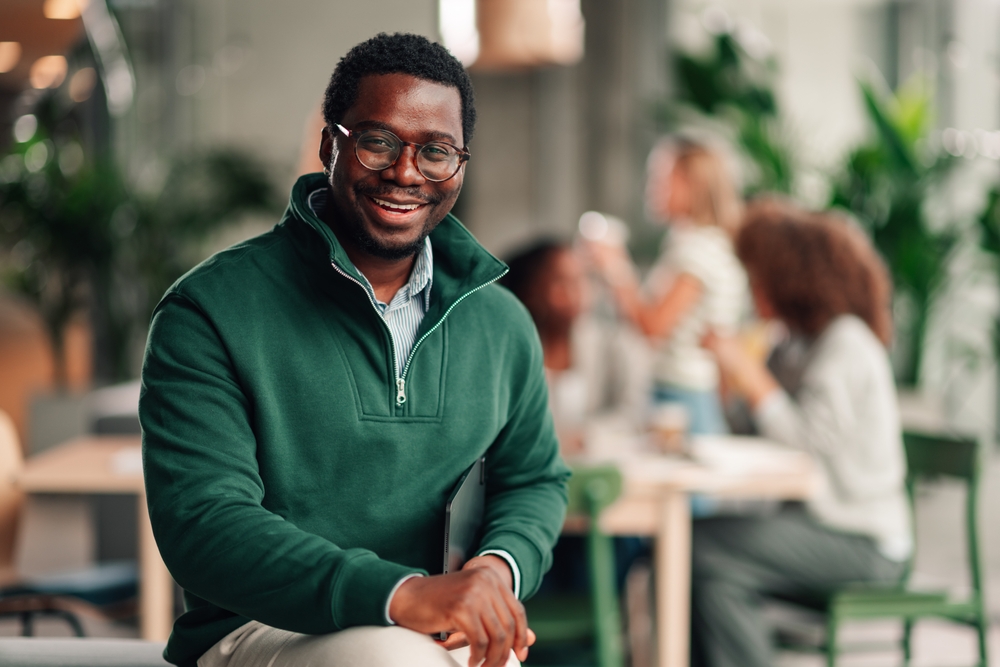 A smiling man wearing glasses and a green pullover seated indoors