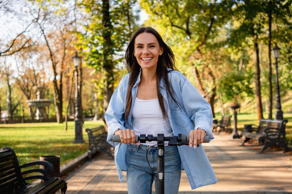 A smiling woman holding the handlebars of a scooter on a tree-lined park path