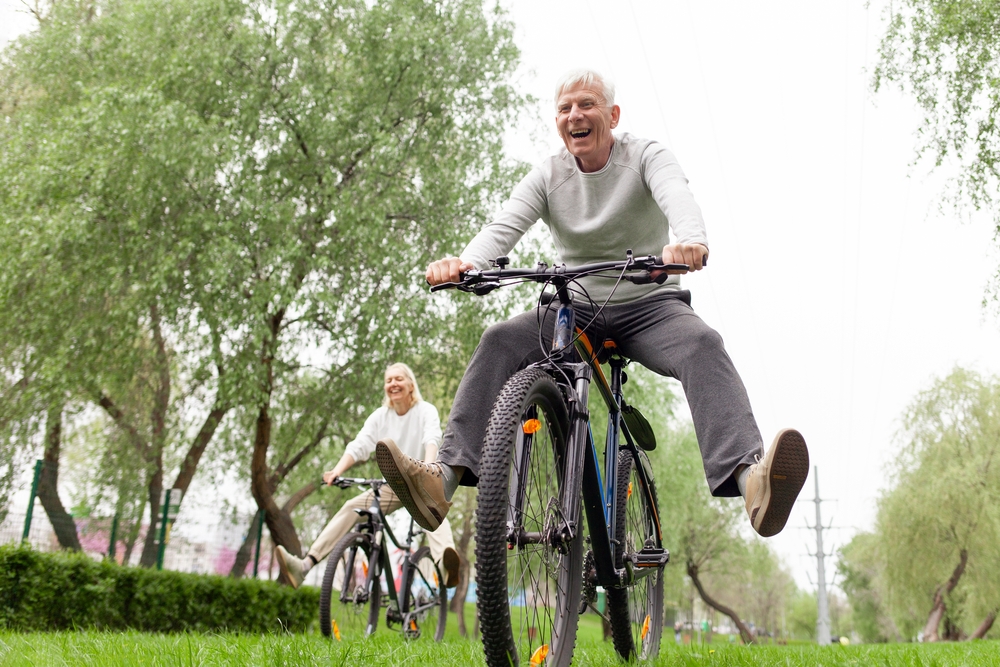 An older couple smiling while riding bicycles along a tree-lined path