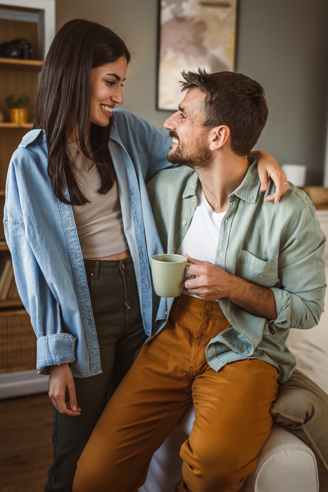 A smiling couple at home, with the woman resting her arm around the man as he holds a mug