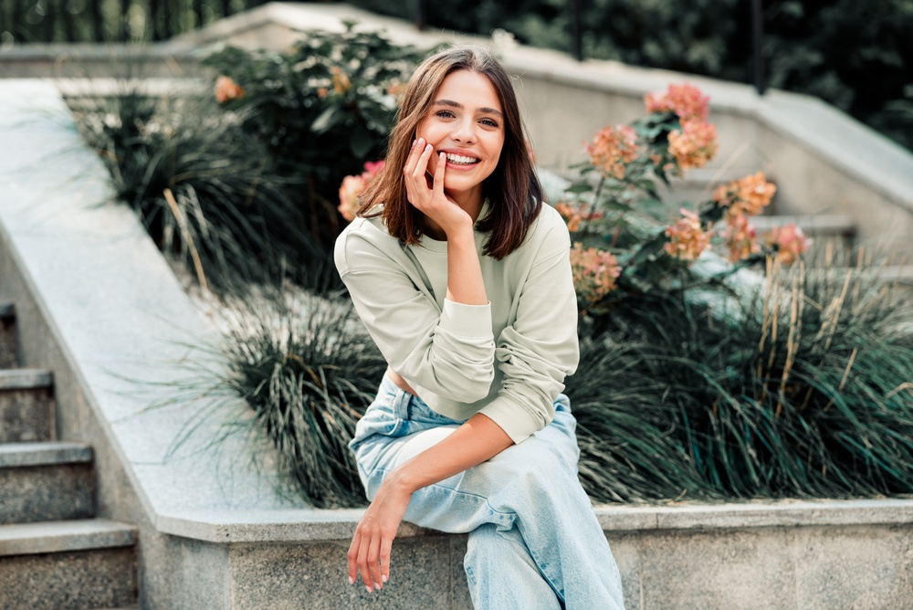 A smiling woman seated beside a garden, resting her hand against her cheek