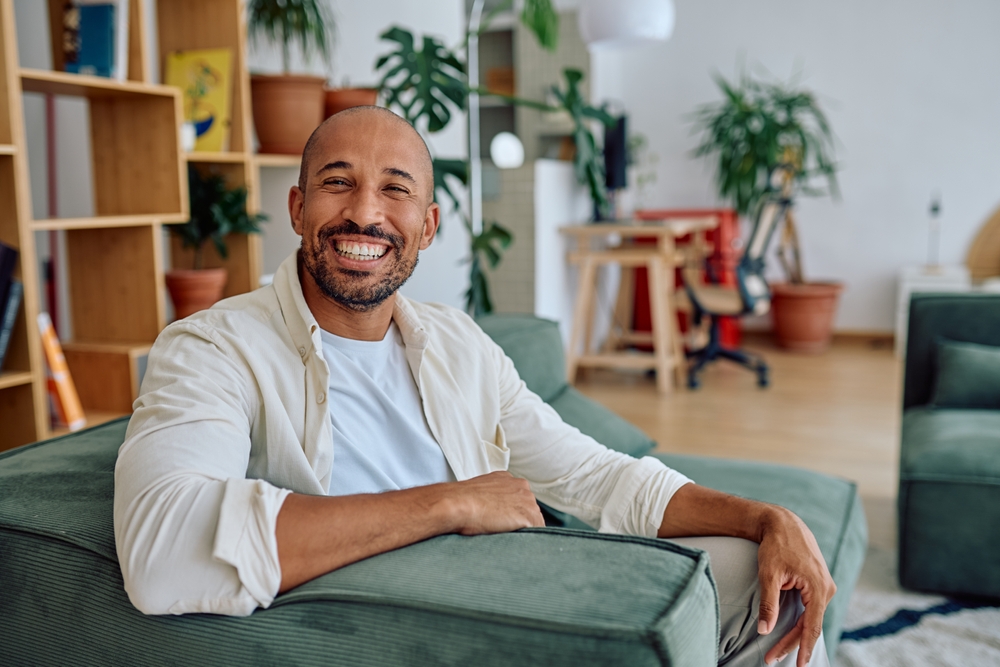 A smiling man relaxing on a green sofa in a bright room with plants