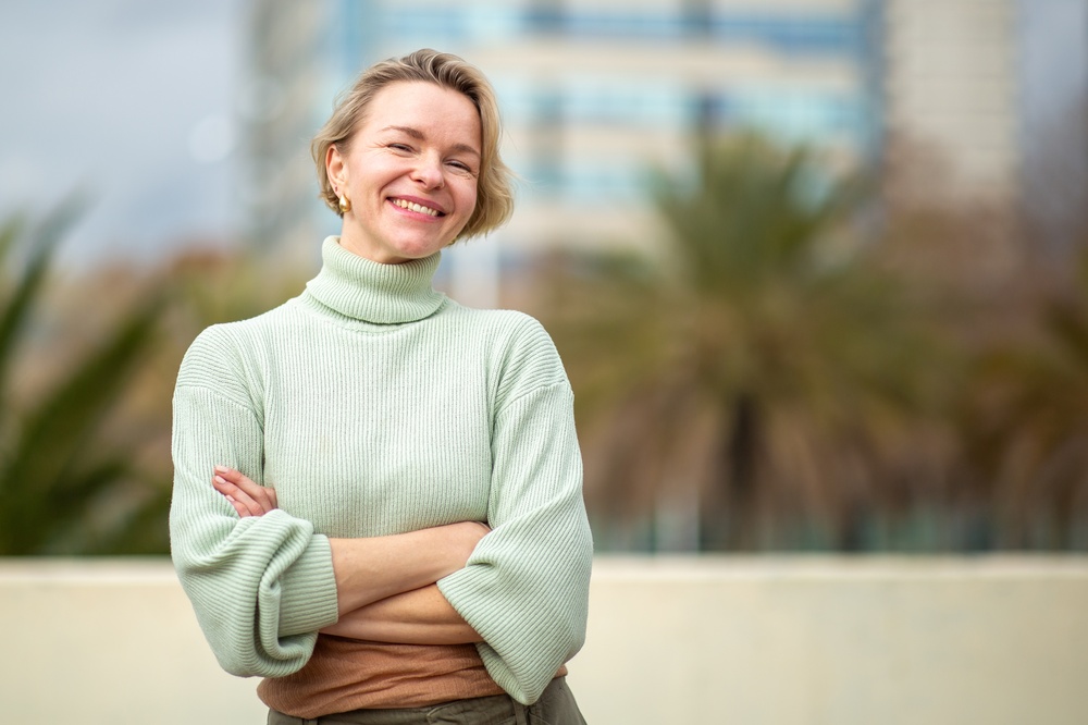 A smiling woman in a pale green sweater standing outdoors with her arms folded