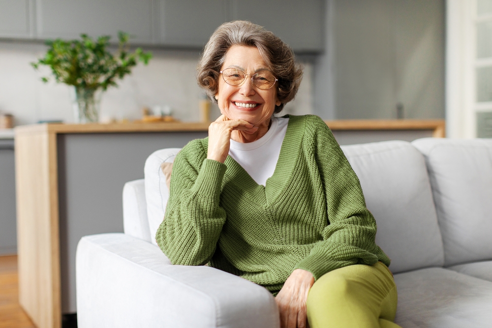 A smiling older woman wearing glasses and a green sweater seated on a sofa