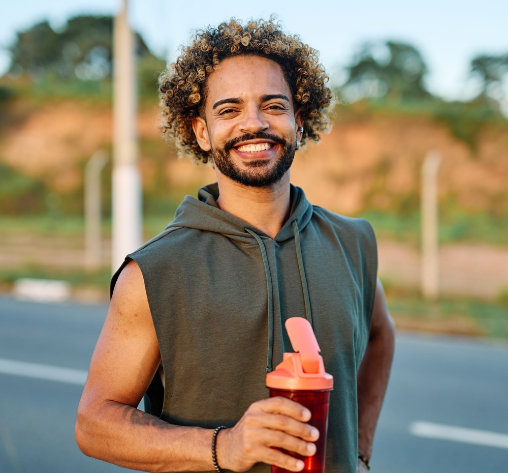 A smiling man in a sleeveless hoodie holding a water bottle outdoors