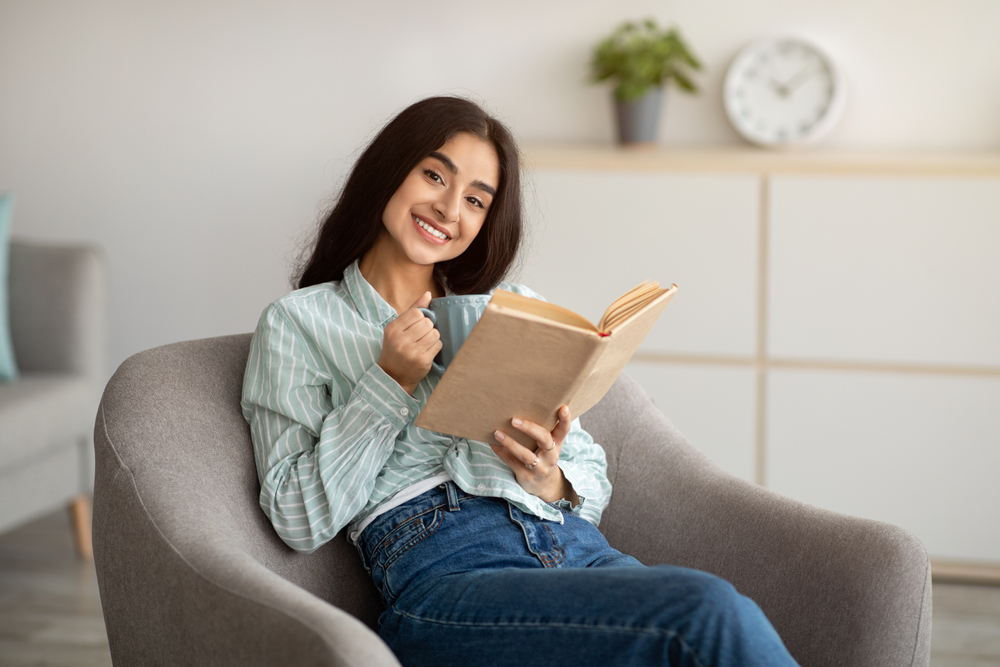 A smiling woman seated in an armchair holding a book and a mug
