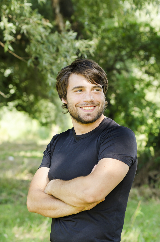 A smiling man in a dark shirt standing outdoors with his arms folded